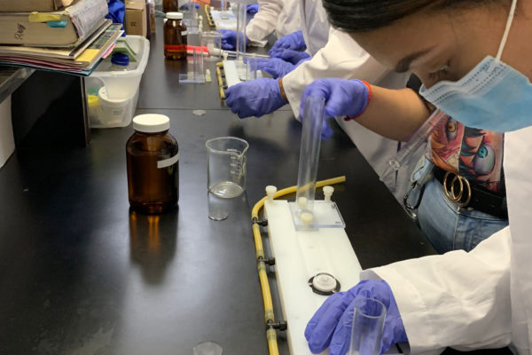 A group of students in lab coats working with beakers and settling chambers while being observed by a mentor on the far end.