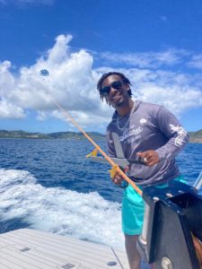 A person on a boat jokingly holds a fake, large pair of scissors up to a rope attached to a person parasailing