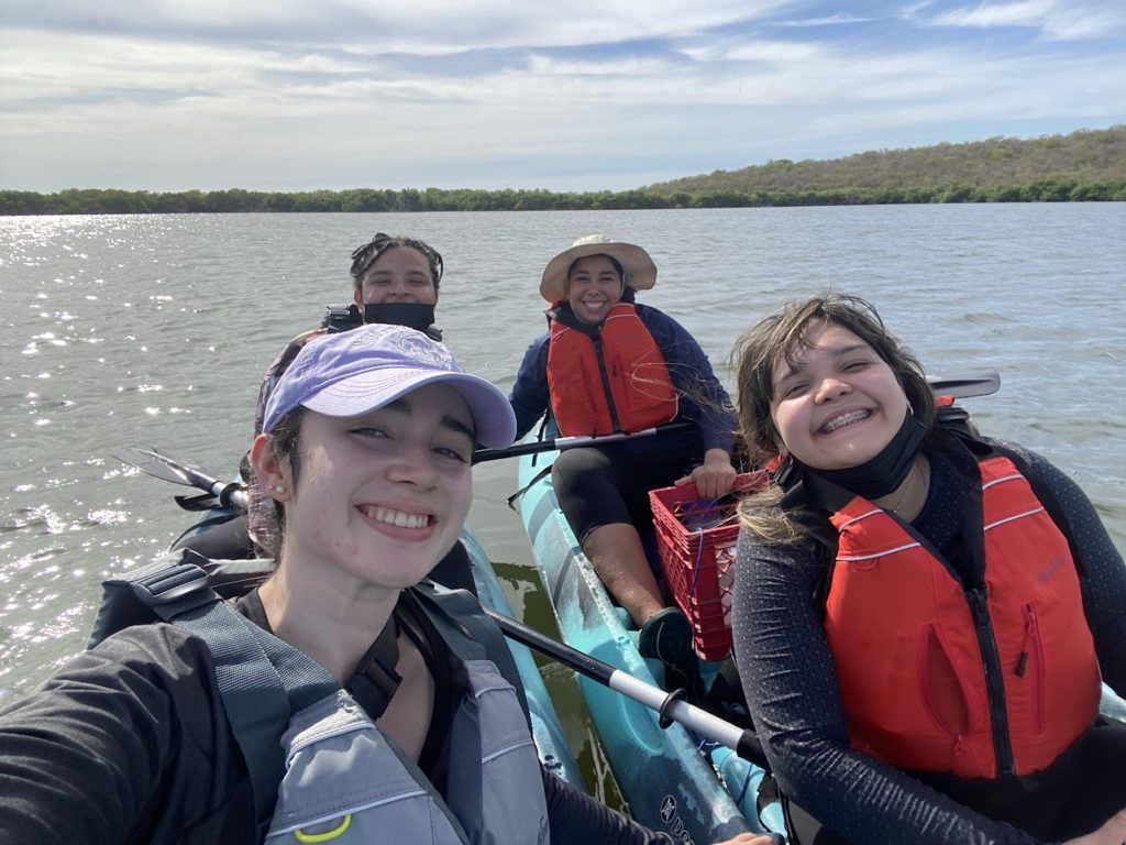Four young women in double kayaks on a body of water. They wear life jackets and smile for a selfie