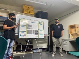 Two students wearing T-shirts, jeans and face masks give a presentation while standing beside a scientific poster